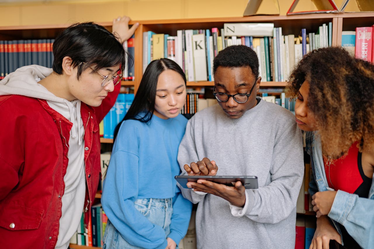 A group of college students collaborate on a tablet in a library surrounded by bookshelves.
